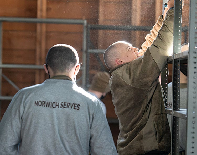 Male cadets. One in a gray "NORWICH SERVES" shirt, the other reaching up to a metal shelf.