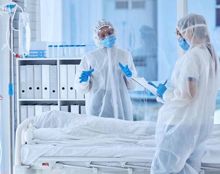 Two healthcare workers in full PPE stand around a patient bed, one talking and the other taking notes.