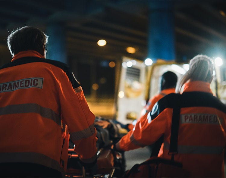 Three paramedics in orange jackets transport a stretcher at night, likely into an ambulance.