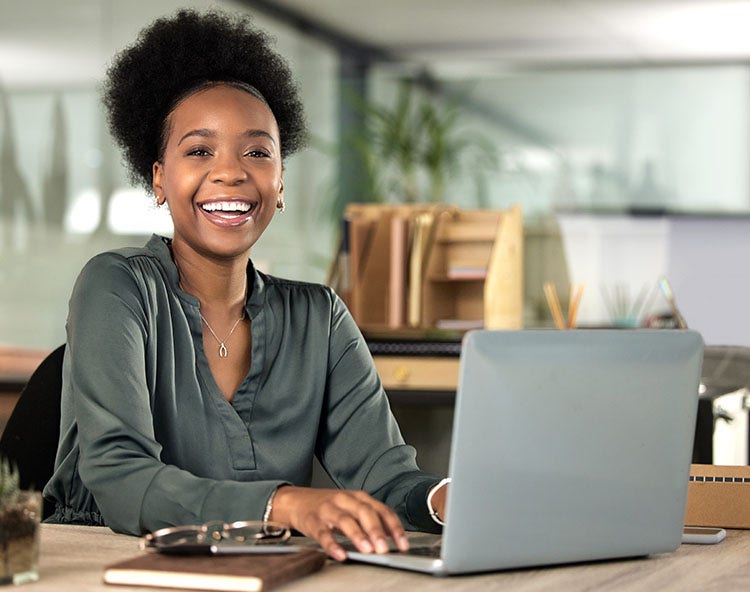 A smiling young Black woman with an afro sits at a desk, typing on a laptop in an office.