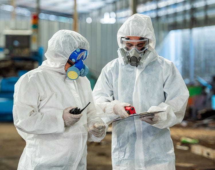 Two people in full-body white hazmat suits and respirators discuss something in an industrial setting.