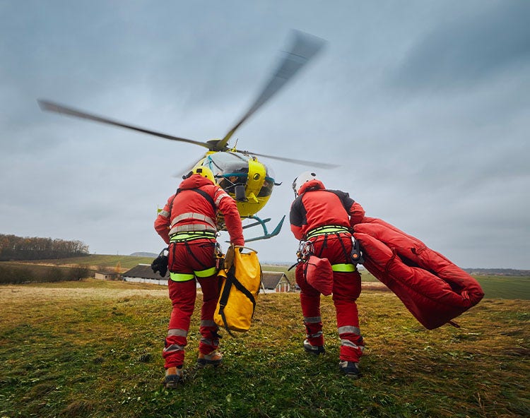 Two rescue workers in red suits and helmets approach a yellow helicopter on a grassy field under a cloudy sky.